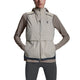 En kvinde står med hænderne på hofterne mod en hvid baggrund, iført en On Weather Vest Dame i Desert/Eclipse over en langærmet taupe top og sorte leggings.
