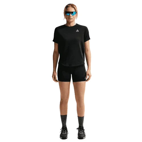 En person står med ansigtet fremad i en sort Summit White Nike ACG Short-Sleeve Running Top (Dame), sorte shorts, grå sokker, sorte sportssko og blå reflekterende solbriller mod en almindelig lys baggrund.