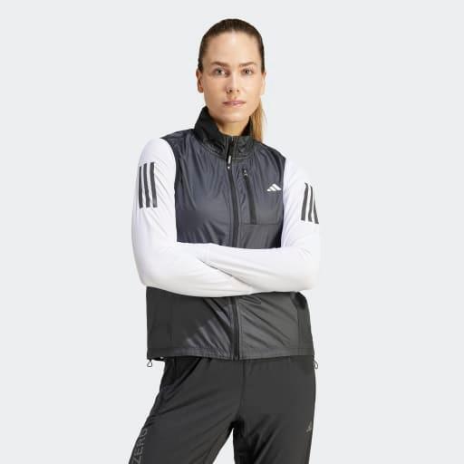En kvinde med langt tilbagestrøget hår bærer Adidas Otr B Vest, Dame, Black over en hvid langærmet skjorte med sorte striber og sorte bukser, mens hun står med armene over kors på en almindelig lys baggrund.