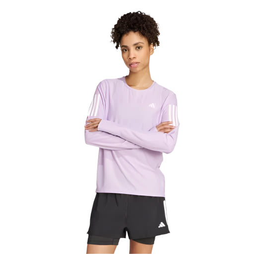 En person er iført en Adidas Otr B Ls, Dame, Powplu løbetrøje i lyslilla med hvide ærmestriber og sorte sportsshorts med hvidt logo, der står på en almindelig hvid baggrund.