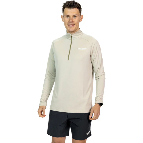 En mand med kort mørkt hår smiler, mens han kigger fremad, i en Liiteguard Ground-Tech 1/4 Zip, Herre, Beige lavet af åndbart materiale og sorte shorts. Trøjen har et lille hvidt logo på venstre bryst.