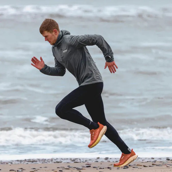 Iført en Liiteguard Aero-Tech Windbreaker til mænd i Dark Shade sammen med sorte leggings og orange løbesko sprinter en person hen over en stenet strand, mens bølgerne slår ind i baggrunden.