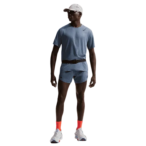 En mand står i et Nike-løbeoutfit - lysegrå kasket og skjorte, AeroSwift 4In Brief-Lined Running Shorts for men i Diffused Blue/Black, knaldorange sokker og hvide løbesko - på en almindelig hvid baggrund.