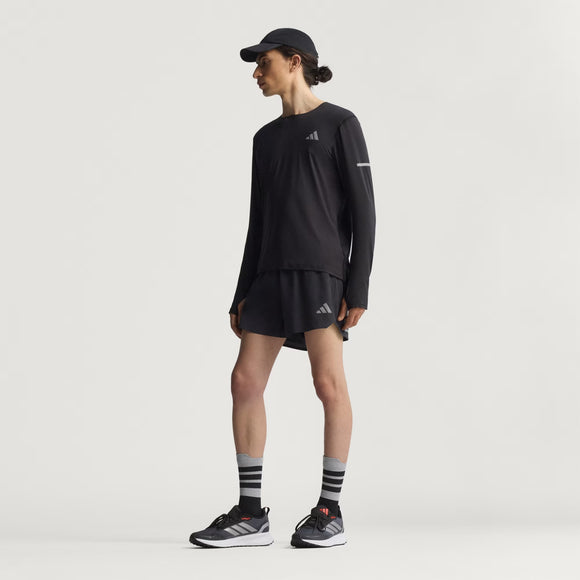 En person står på en ensfarvet baggrund iført en sort langærmet skjorte, Adidas Adi365 Short M Herre i sort, en sort kasket, sorte sneakers med hvide såler og sort-hvid-stribede sokker.