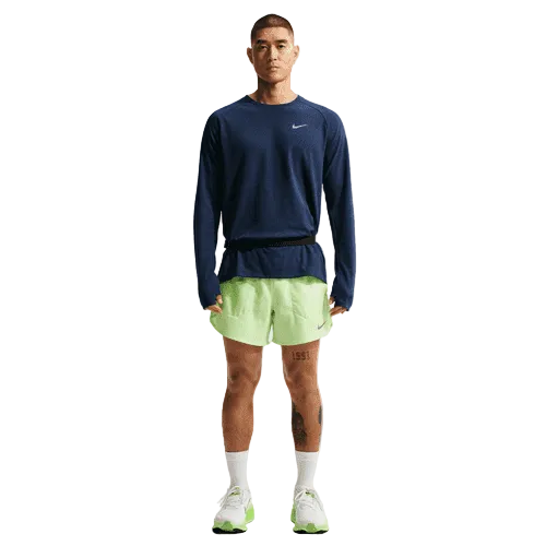 En person står og kigger fremad iført en Nike Stride Waffle Crew-løbetrøje til mænd i Midnight Navy/Reflective Silver, lysegrønne Nike-shorts, hvide sokker og lysegrønne løbesko med reflekterende detaljer på en almindelig hvid baggrund.