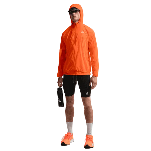En person står på en ensfarvet baggrund iført en orange jakke med hætte, sorte shorts, Nike ACG Lavaloops 1/2 Tights Herre Black/Summit White, orange løbesko, hvide sokker, en sort kasket, solbriller og med en sort vandflaske i hånden - ideel træningstøj.