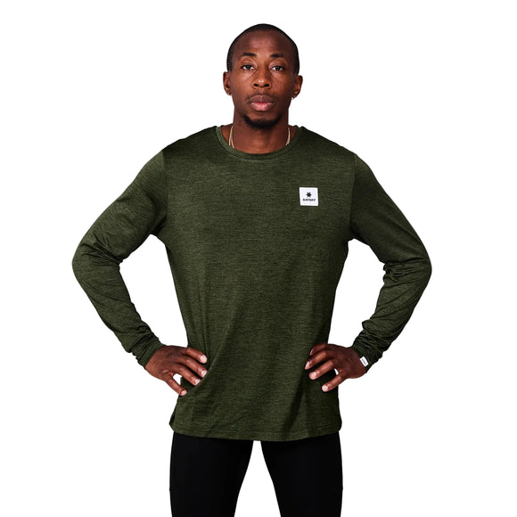 En mand står på en almindelig grå baggrund iført Saysky Clean Pace Long Sleeve, Herre, Green med et lille hvidt mærke på brystet og sorte bukser. Hans hænder er på hofterne, mens han ser direkte ind i kameraet.