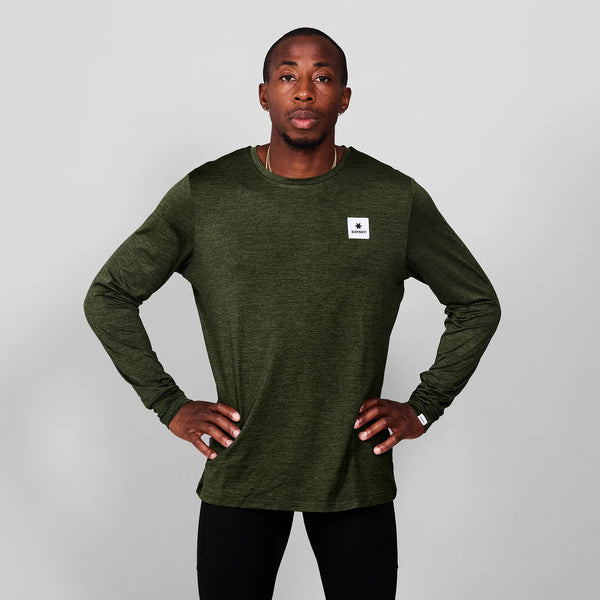 En mand står på en almindelig grå baggrund iført Saysky Clean Pace Long Sleeve, Herre, Green med et lille hvidt mærke på brystet og sorte bukser. Hans hænder er på hofterne, mens han ser direkte ind i kameraet.