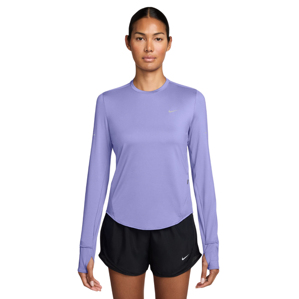 En person står med ansigtet vendt fremad og har en Nike Swift UV langærmet løbetrøje til kvinder i farven Purple Pulse/Reflective Silver på, kombineret med sorte Nike-shorts. Baggrunden er ensfarvet hvid, og personen har et neutralt udtryk med armene afslappet ned langs siden.