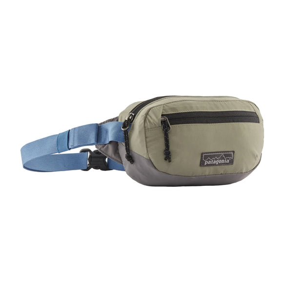 Patagonia Terravia Mini Hip Pack, Unisex, i River Rock Green har et olivengrønt og gråt design med en sort lynlåslomme foran, blå justerbar rem og sort spænde til kompakt opbevaring, vist på en hvid baggrund.
