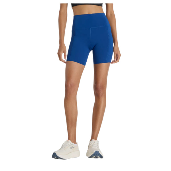 En person står på en hvid baggrund, hvor man kun kan se underkroppen og benene, iført New Balance Sleek Pocket High Rise Short 6" i Galaxy Blue og hvide sportssko.