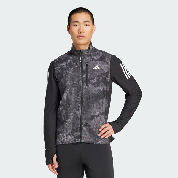 En person med kort hår bærer en sort og grå mønstret Adidas-jakke med lynlås og Adidas Otr Sd Vest M til mænd i Grefou/Black, mens han står med en hånd på hoften mod en almindelig lys baggrund.