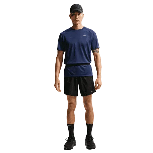 En person står med ansigtet fremad og er iført en Nike Stride SS løbetop til mænd i Midnight Navy/Reflective Silver sammen med sorte shorts, strømper, sportssko og en sort kasket. Baggrunden er helt hvid.