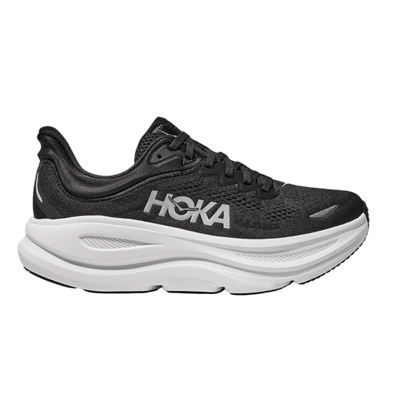 Hoka Bondi 9 Dame i sort/hvid har en overdel i mesh og en tyk hvid sål med »HOKA« trykt i hvidt på siden. Denne sportslige løbesko, der her ses set fra siden på en ensfarvet hvid baggrund, er designet til maksimal komfort.