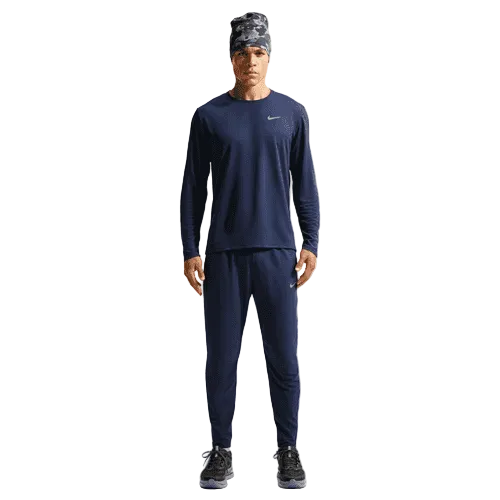 En person står og kigger fremad iført en marineblå langærmet Nike-trøje, Nike Stride Woven løbebukser til mænd i Midnight Navy/Reflective Silv, sorte løbesko og en mønstret hue på en ensfarvet hvid baggrund.