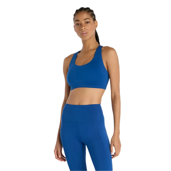 En kvinde med flettet hår er iført New Balance Define Train Bra, Dame, i Galaxy Blue, sammen med matchende højtaljede leggings, der står på en almindelig hvid baggrund.
