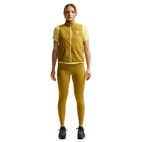 En kvinde står og kigger fremad iført Nike ACG 7/8 Running Tights i Peat Moss/Ironstone/Summit White med en matchende ærmeløs vest med lynlås og sorte sneakers på en almindelig hvid baggrund.