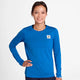 En kvinde med brunt hår bundet tilbage bærer den lyseblå Saysky W Clean Pace Long Sleeve, Dame, med et lille hvidt rektangulært logo på brystet. Hun smiler til kameraet mod en almindelig hvid baggrund.