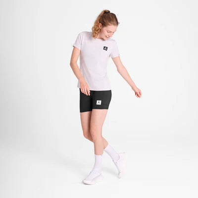 En kvinde står på en almindelig hvid baggrund iført Saysky W Statement Pace T-shirt i hvid, sorte shorts, hvide sokker og hvide sneakers. Hun kigger ned og smiler med armene lidt ud til siderne.