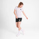 En kvinde står på en almindelig hvid baggrund iført Saysky W Statement Pace T-shirt i hvid, sorte shorts, hvide sokker og hvide sneakers. Hun kigger ned og smiler med armene lidt ud til siderne.