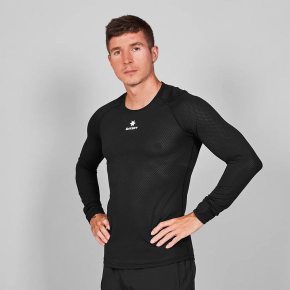 En mand med kort brunt hår viser Saysky Mesh Base Layer Long Sleeve, Unisex, i sort med matchende bukser. Han står på en almindelig grå baggrund med hænderne på hofterne og kigger direkte ind i kameraet.