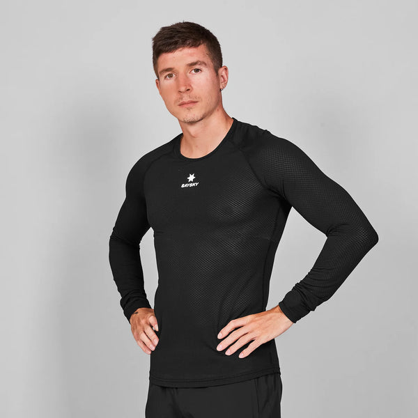 En mand med kort brunt hår viser Saysky Mesh Base Layer Long Sleeve, Unisex, i sort med matchende bukser. Han står på en almindelig grå baggrund med hænderne på hofterne og kigger direkte ind i kameraet.