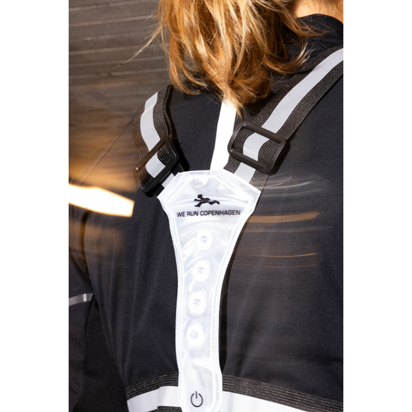 En person med langt hår bærer en sort jakke og BeeSafe LED Harness USB Phone Pocket, Unisex, Silver - ideel til natløb. På vesten står der "WE RUN COPENHAGEN" med fire knapper. Baggrunden er sløret og stærkt oplyst indendørs.