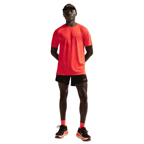 En person står med ansigtet fremad i en knaldrød kortærmet sportstrøje, Nike Stride 5" Brief-Lined Running Shorts til mænd i Black/Reflective Silver, sorte og orange Nike-løbesko, røde sokker, en sort kasket og en almindelig hvid baggrund.