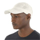 En mand iført en Salomon SHKOUT Cap U, Whisper White - en hvid hurtigtørrende kasket med små ventilationshuller og justerbar pasform - og en mørk crew neck trøje kigger let til venstre mod et ensfarvet hvidt baggrund.