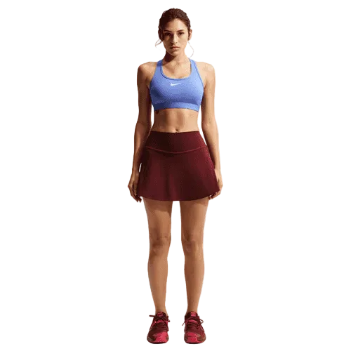 En kvinde iført en blå Nike Swoosh Medium Sports Bra (Dame, Sapphire/White), en bordeaux sportskørt samt matchende sneakers står på en ensfarvet hvid baggrund.