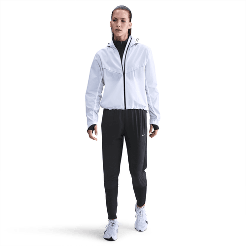 En person står og kigger fremad iført en hvid jakke med lynlås, Nike Dri-FIT Mid-Rise Running Pants for Dame i Black/Reflective Silv og hvide sportssko, der viser stilfuld træningstøj dame mod en almindelig hvid baggrund.