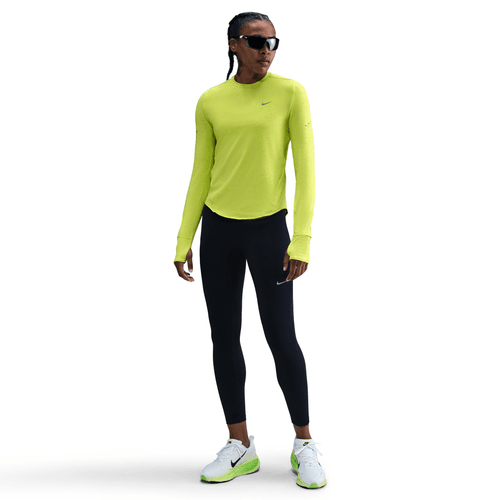 En person iført en Nike Swift UV Long Sleeve Shirt til kvinder i Cyber/Htr/Reflective Silver, sorte leggings, hvide sportssko med grønne detaljer og solbriller står på en almindelig hvid baggrund.