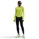 En kvinde iført en neongul Nike Dri-FIT UV Long-Sleeve Crew-Neck Running Top (Dame, Cyber/Htr/Reflective Silv), sorte leggings, hvide og grønne sportssko og sorte solbriller står på en almindelig hvid baggrund.
