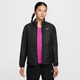 En kvinde med langt flettet hår er iført en sort Nike Therma-FIT løbejakke (Dame, Black/Reflective Silv) over en lyserød skjorte og sorte bukser, mens hun står på en almindelig hvid baggrund.