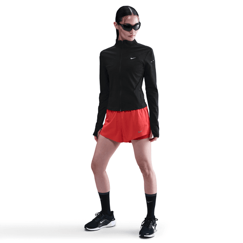 På en hvid baggrund bærer en person en sort Nike Dri-FIT Full-Zip Running Mid Layer til kvinder i Black/Reflective Silv, sammen med røde shorts, sorte strømper, sorte løbesko og store sorte solbriller.