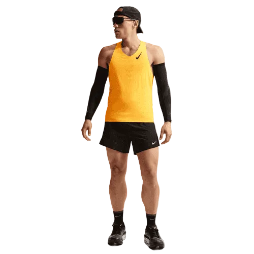 En person i Nike AeroSwift Running Singlet, Herre, Laser Orange/Black, sorte løbeshorts, ærmer, sko, kasket og solbriller står vendt lidt til siden mod en ensfarvet baggrund.