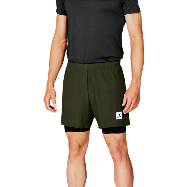 En person iført en sort kortærmet skjorte og Saysky 2 in 1 Pace Shorts 5'', Herre, Green med et lille rektangulært mærke på venstre ben, står på en hvid baggrund.