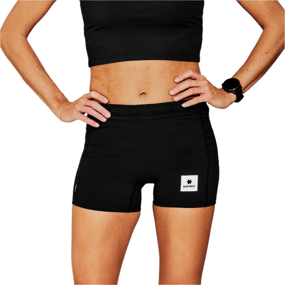 En person står med hænderne på hofterne iført en sort crop top og Saysky W Flow+ Race Short Tights 4" til kvinder i sort. Et hvidt mærke er synligt på venstre ben, og et sort armbåndsur bæres på venstre håndled. Baggrunden er hvid.