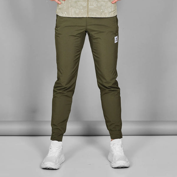 En mand iført Saysky Pace Pants, Herre, Green med en lys mønstret skjorte og hvide sportssko står på en almindelig grå baggrund. Billedet er beskåret ved skuldrene.