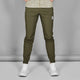 En mand iført Saysky Pace Pants, Herre, Green med en lys mønstret skjorte og hvide sportssko står på en almindelig grå baggrund. Billedet er beskåret ved skuldrene.