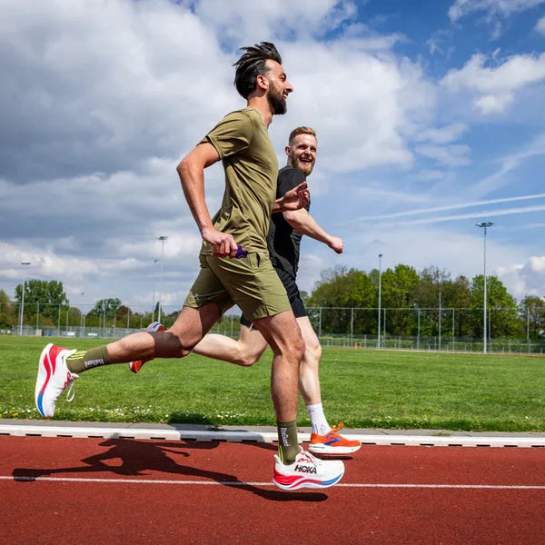 To mænd løber udendørs under en delvis overskyet himmel. Den ene er iført LiiteGuard Ground-Tech T-shirt, Herre, i Dusty Green. De er klædt i sportstøj og sneakers, og i baggrunden ses græs, træer og lysmaster.