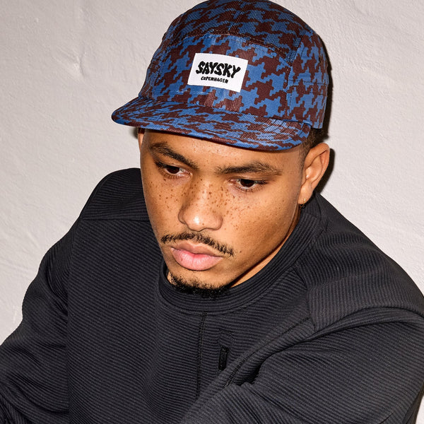En mand med fregner og kort skæg bærer Saysky Houndstooth Combat Cap 101, Unisex, Blue Aop med Copenhagen-logo og en sort tekstureret sweatshirt, mens han kigger ned mod en ensfarvet lys væg.
