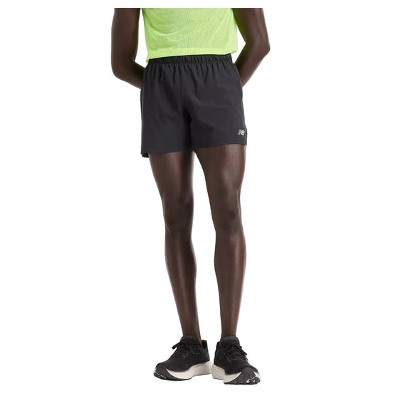 En person står med hænderne på ryggen, vist fra taljen og ned, iført en neongrøn skjorte, sorte New Balance RC Short 5" Herre-shorts og sorte løbesko med hvide såler på en almindelig hvid baggrund.