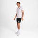 En person står i et almindeligt hvidt rum iført en hvid Saysky Statement Pace T-shirt i hurtigtørrende materiale, sorte shorts, hvide sokker og hvide sneakers. Både trøjen og shortsene har et lille Saysky-logo på forsiden.