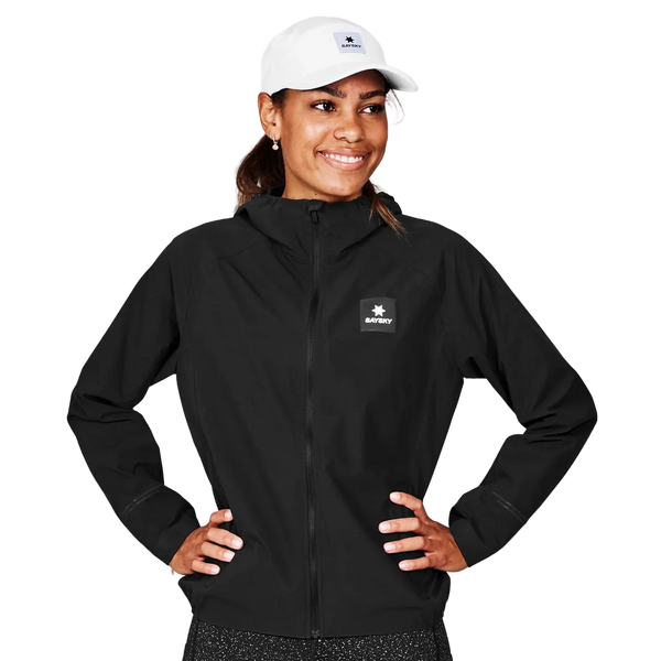En kvinde står med hænderne på hofterne iført en sort Saysky Clean Pace Flex Hooded Jacket, Dame og en hvid baseballkasket. Træningsjakke kvinder har et lille logo på venstre bryst. Hun smiler og kigger lidt til siden.