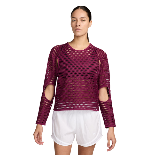 En person har en mørk bærfarvet Nike Aero-FIT langærmet løbetrøje til kvinder, True Berry/Black, med udskæringer ved albuerne og skuldrene, på over en sort sports-BH, kombineret med hvide sportsshorts.
