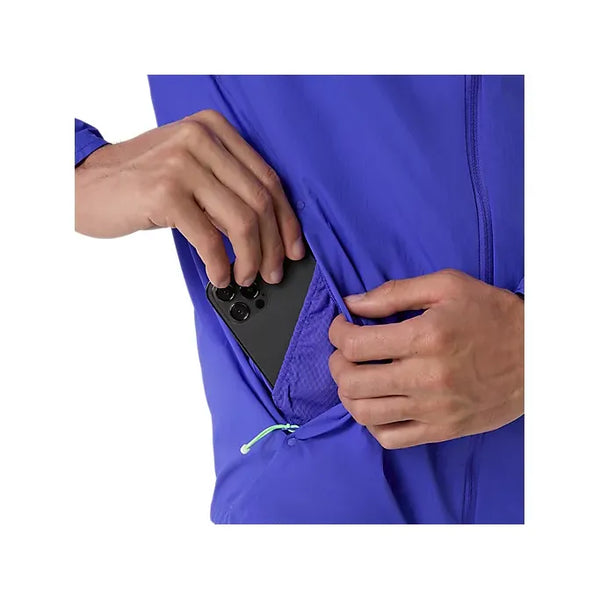 En person iført en Asics Road Packable Jacket, Herre, Cobalt Burst/Illuminate Green lægger en sort smartphone i jakkens sidelomme med lynlås.