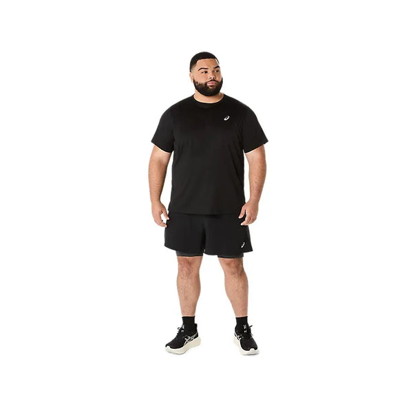 En mand står på en hvid baggrund iført Asics Road 2-N-1 5In Short, Herre, i Performance Black/Graphite Grey, en sort kortærmet atletisk skjorte, sorte sokker og atletiske sko. Han kigger lidt til siden med afslappede arme.