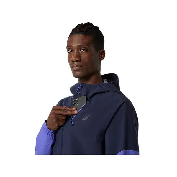 En person iført ASICS Metarun Legend Jacket, Herre, i Midnight/Cobalt Burst/Illuminate Green holder en smartphone tæt på brystet mod en almindelig hvid baggrund.