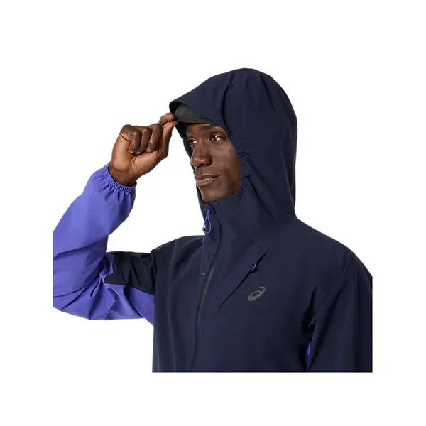 En mand justerer hætten på ASICS Metarun Legend Jacket til mænd i Midnight/Cobalt Burst/Illuminate Green. Denne vandtætte jakke har en lynlås foran og et lille logo på brystet, vist mod en almindelig hvid baggrund.